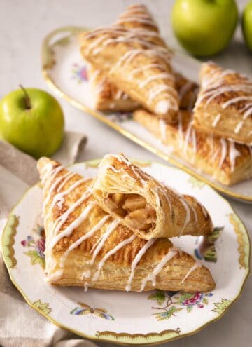 Two apple turnovers on a porcelain plate next to green apples. A bite taken out of a turnover on top.