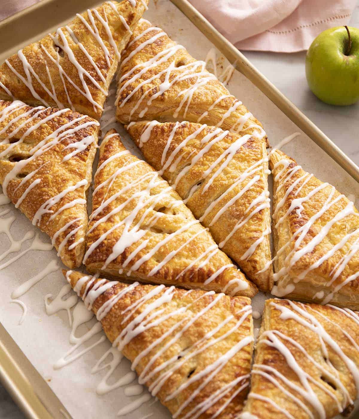 Apple turnovers arranged on a baking sheet. Glaze drizzled on top.