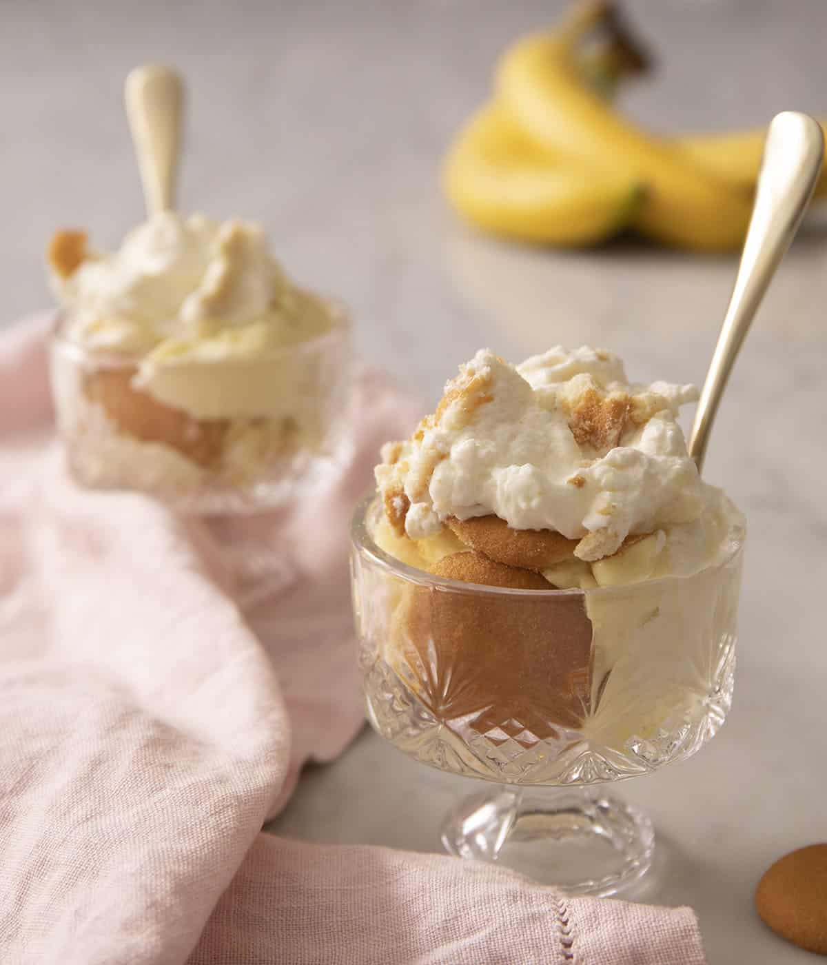 Two servings of banana pudding in crystal glasses with spoons. Bananas in the background.