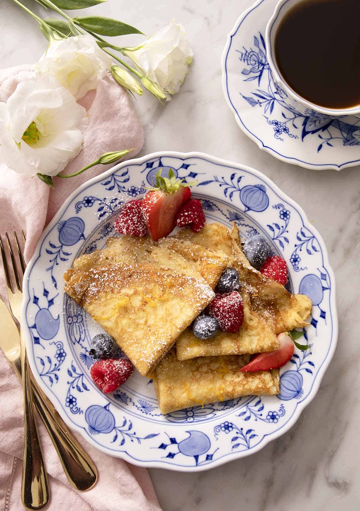 Folded crepes with berries and powdered sugar on a blue and white plate.