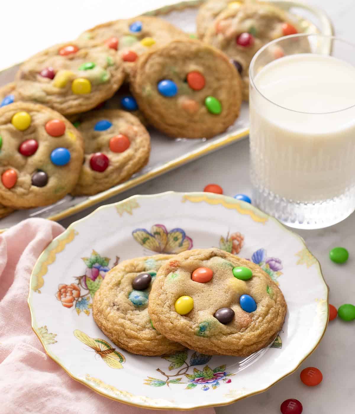 Two M&M cookies on a plate next to a glass of milk. A platter of more cookies in the back. 