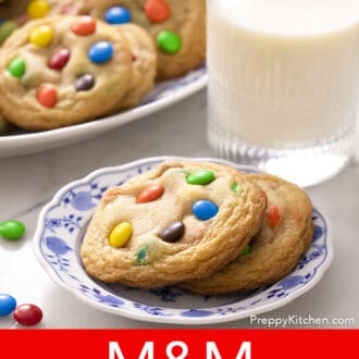 Pinterest graphic of two M&M Cookies on a blue and white plate with a glass of milk and platter in the back.