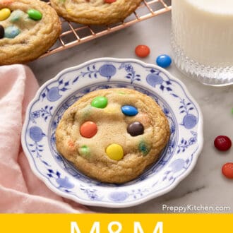 Pinterest graphic of a M&M Cookie on a blue and white plate with a glass of milk and cooling rack of cookies.
