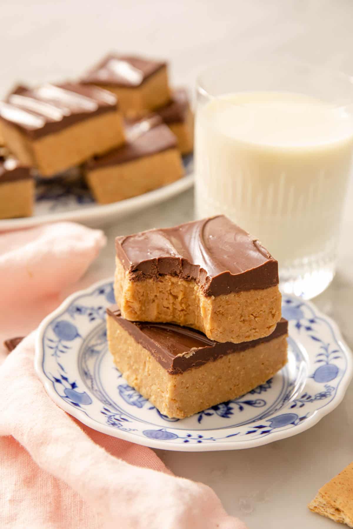 Two peanut butter bars stacked on top of each other on a small plate next to a glass of milk. More bars in the background.