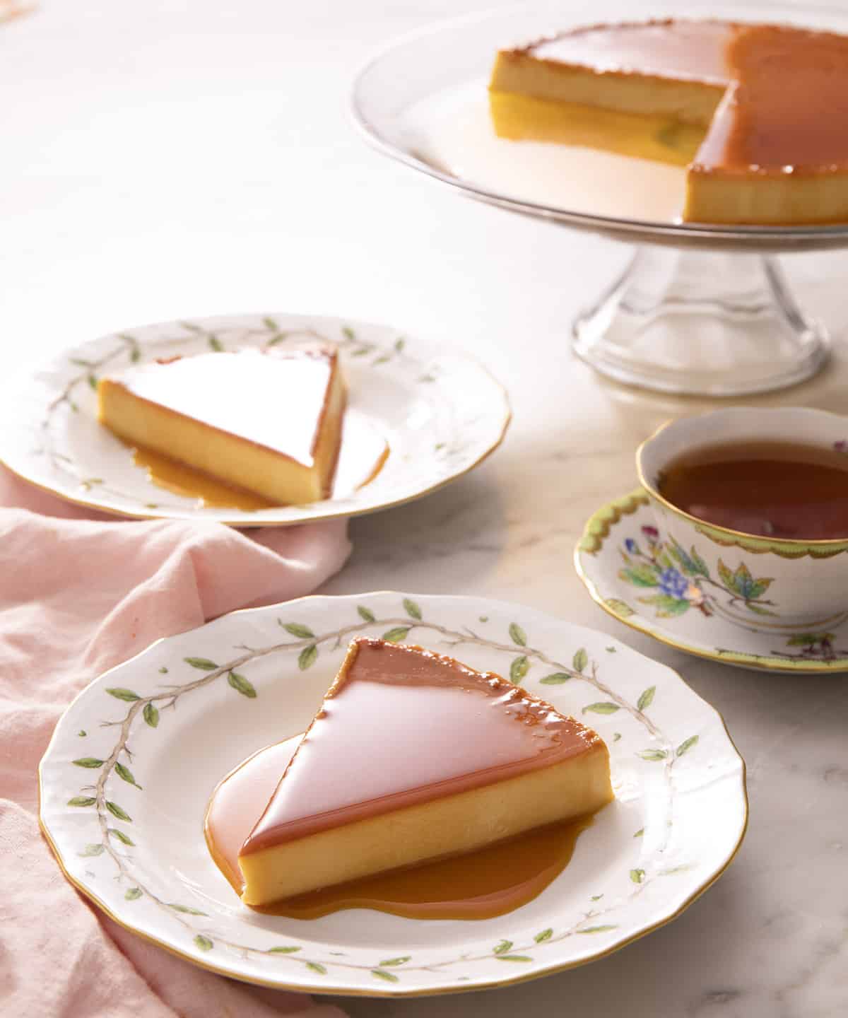 Pieces of flan on porcelain plates next to a cup of tea and the rest of the flan on a cake stand.