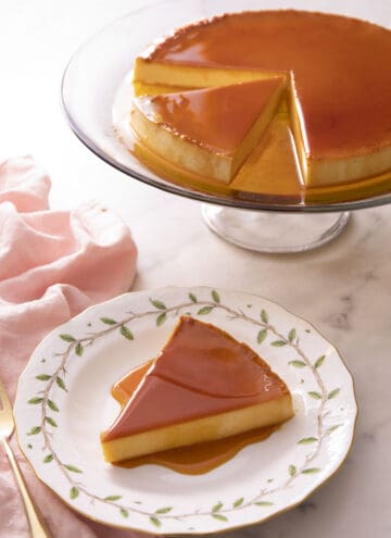A slice of flan on a plate with the rest of the flan on a stand in the background.