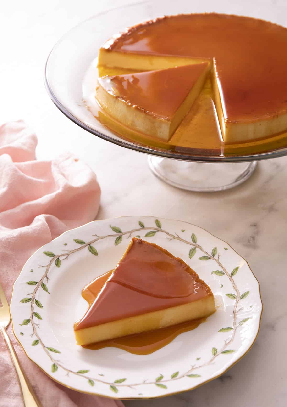 A slice of flan on a plate with the rest of the flan on a stand in the background.