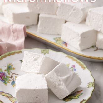 Pinterest graphic of three cubed marshmallows stacked on a floral plate with more marshmallows on a platter in the background.