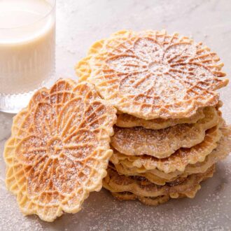 A close up of a stack of pizzelle cookies with a glass of milk in the background