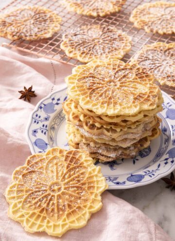 A plate with a stack of pizzelle cookies with more in the background on a cooling rack and one leaning on the plate.