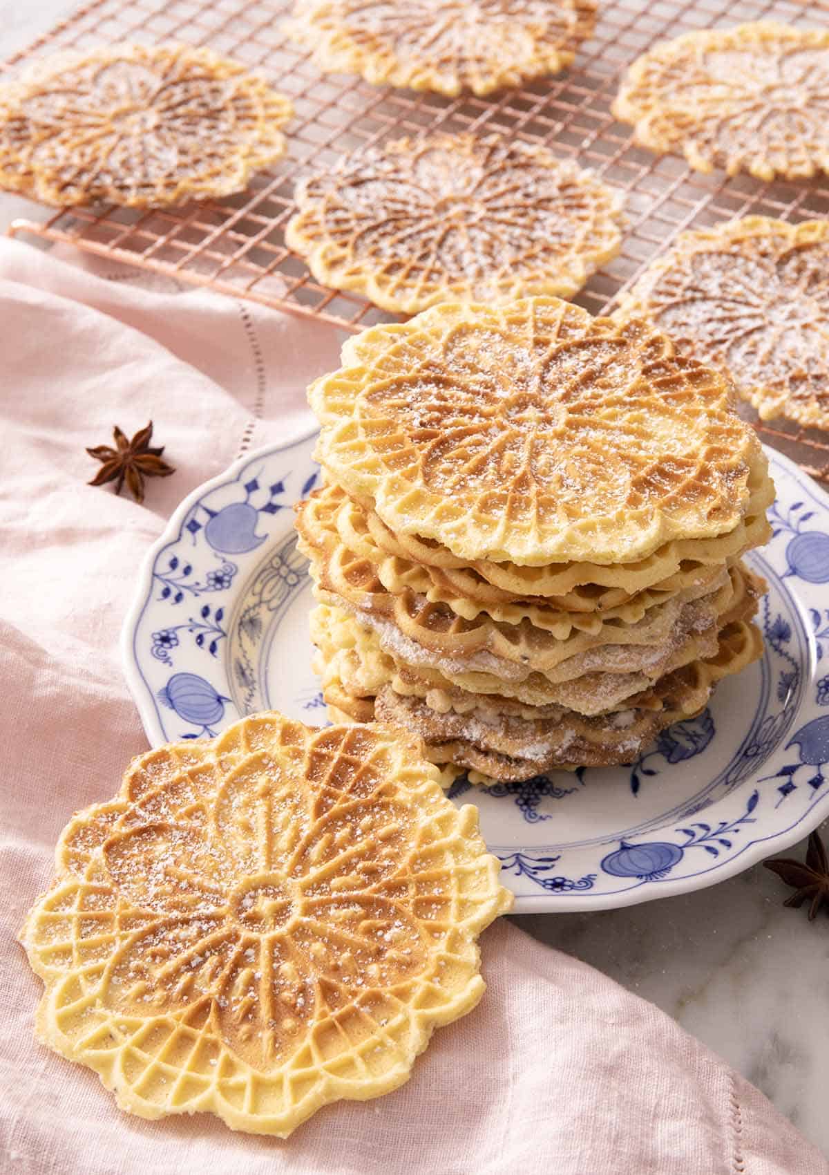 A plate with a stack of pizzelle cookies with more in the background on a cooling rack and one leaning on the plate.