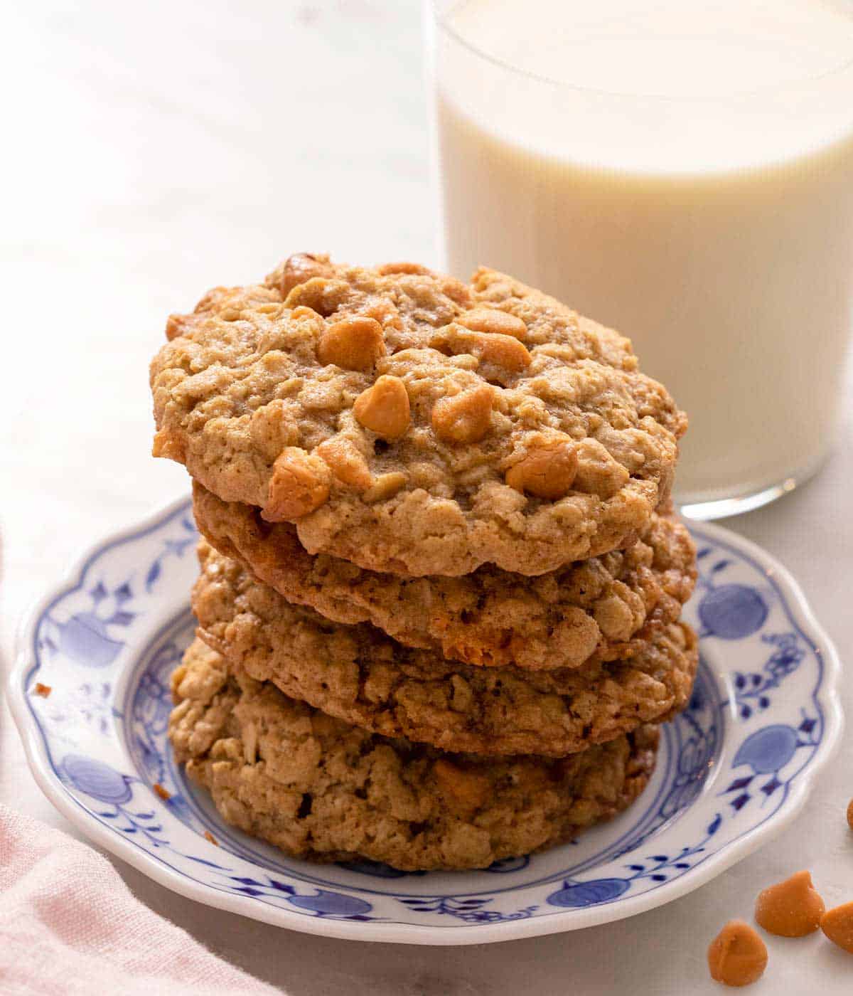 Oatmeal scotchies stacked up on a plate with a glass of milk behind it.