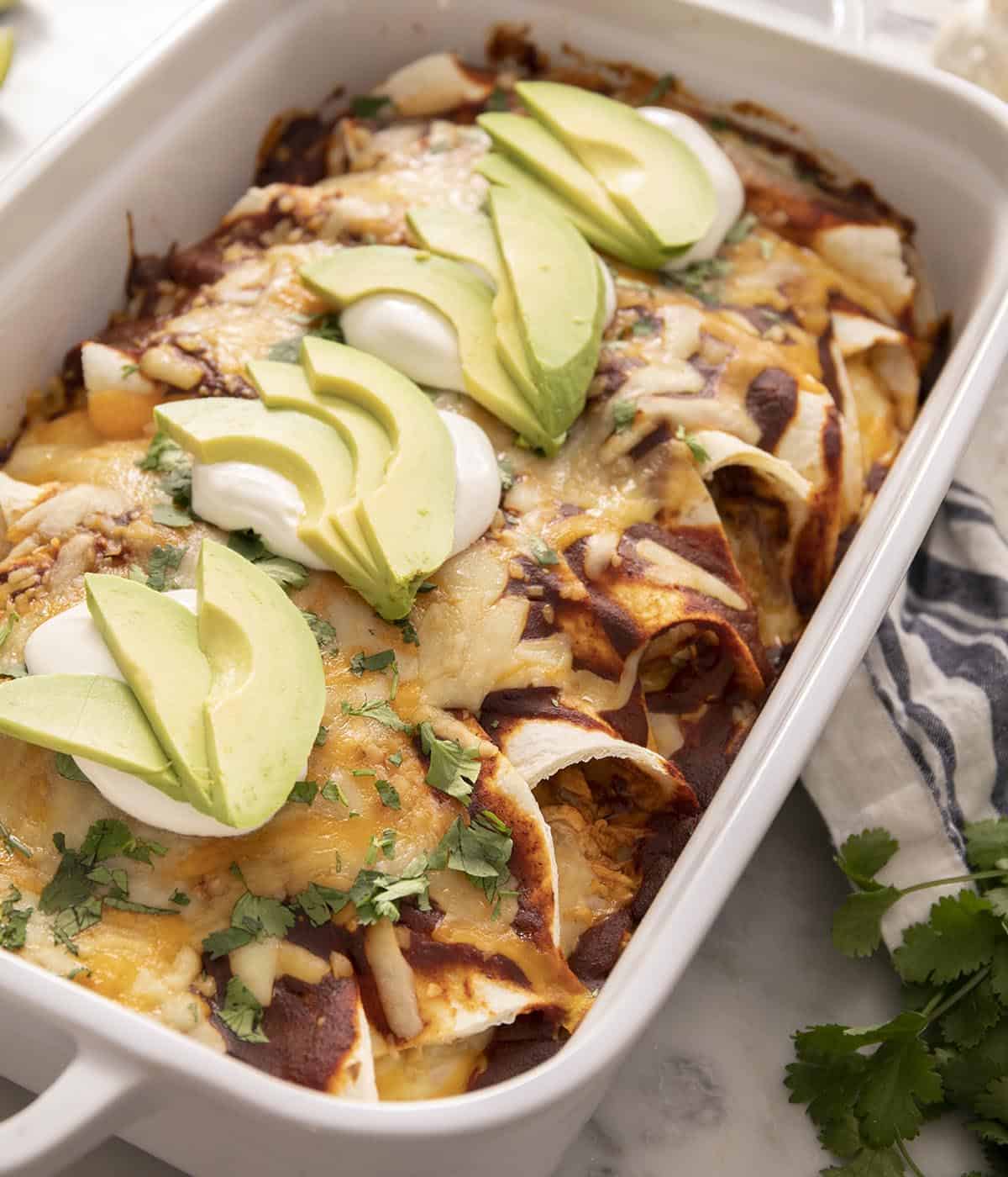 Enchiladas in a casserole dish topped with sour cream and sliced avocado.