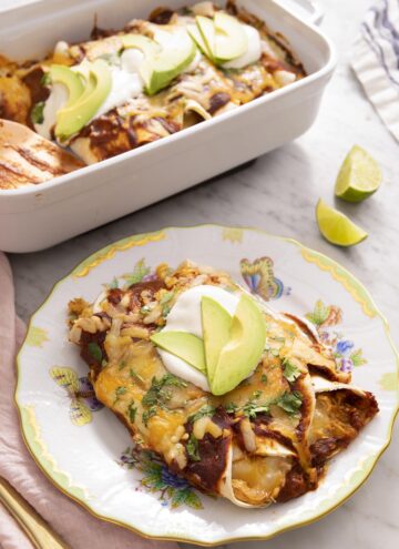 Chicken enchiladas on a plate with sour cream and avocado on top, with a casserole dish of enchiladas in the background.