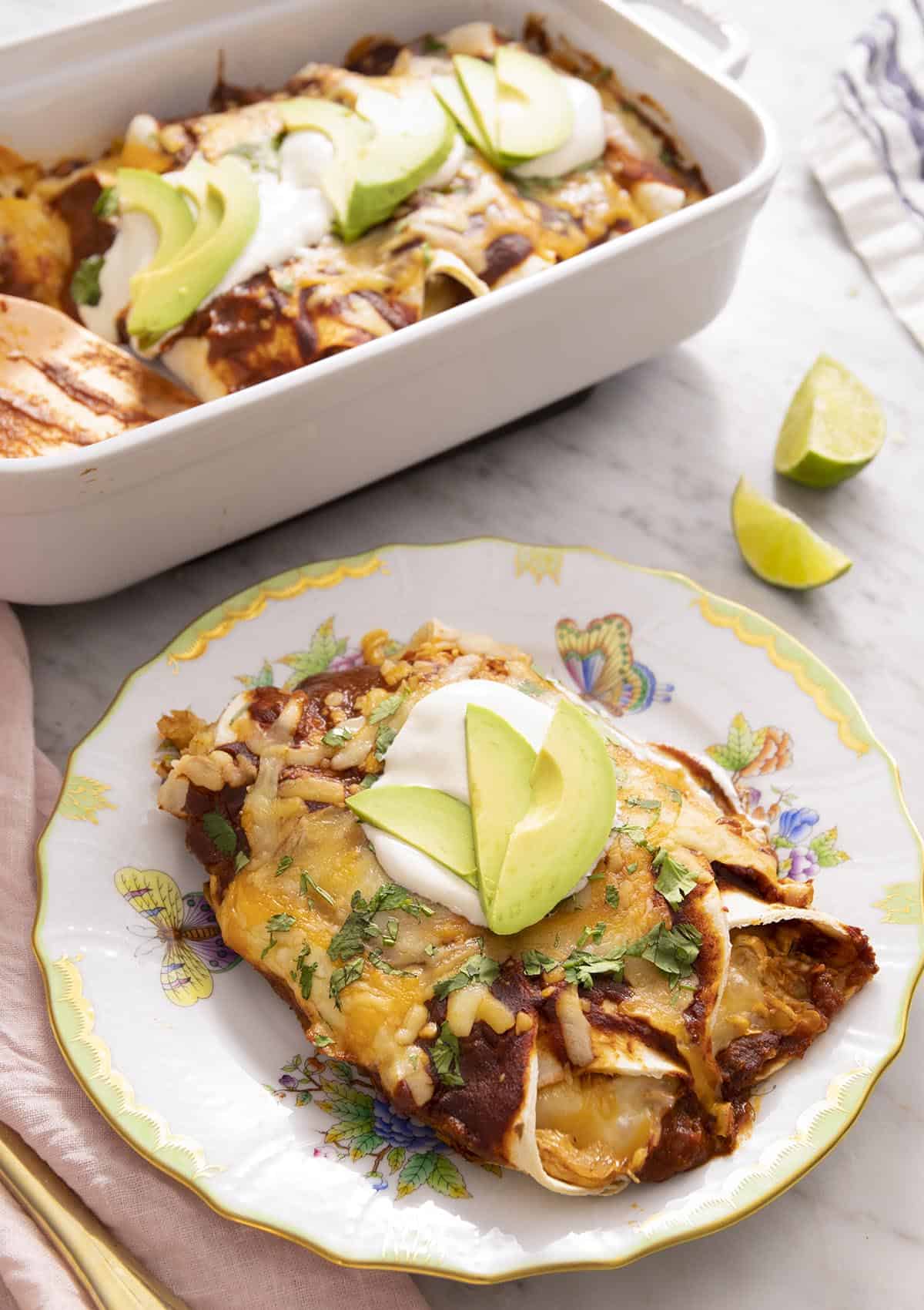 Chicken enchiladas on a plate with sour cream and avocado on top, with a casserole dish of enchiladas in the background.