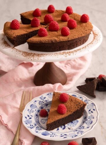 A flourless chocolate cake on a cake stand with a slice on a plate.