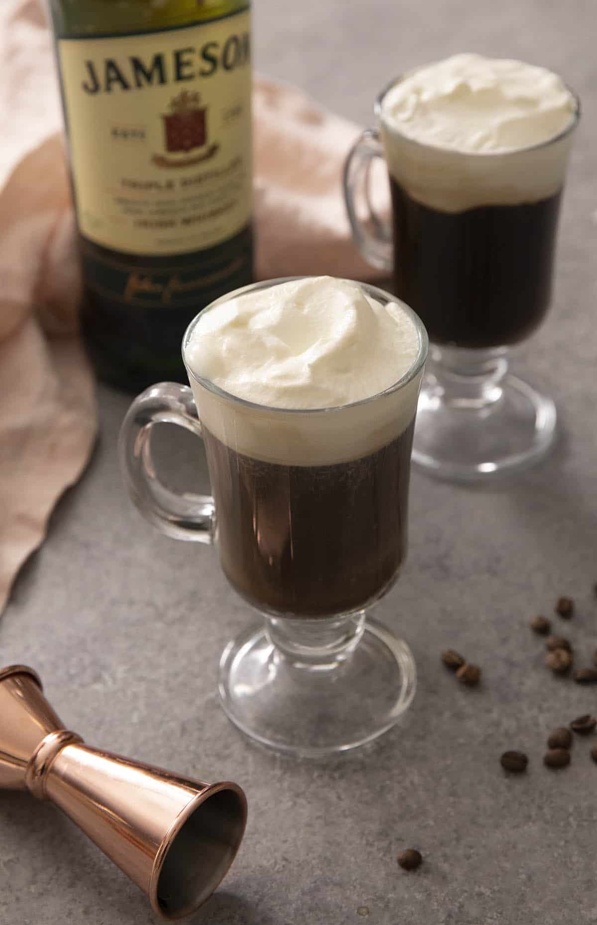 Two mugs on Irish coffee on a work surface with a bottle of whiskey in the background