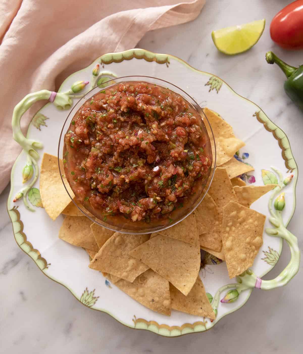 An overhead shot of salsa in a bowl with a bunch of tortilla chips on a plate beside it.