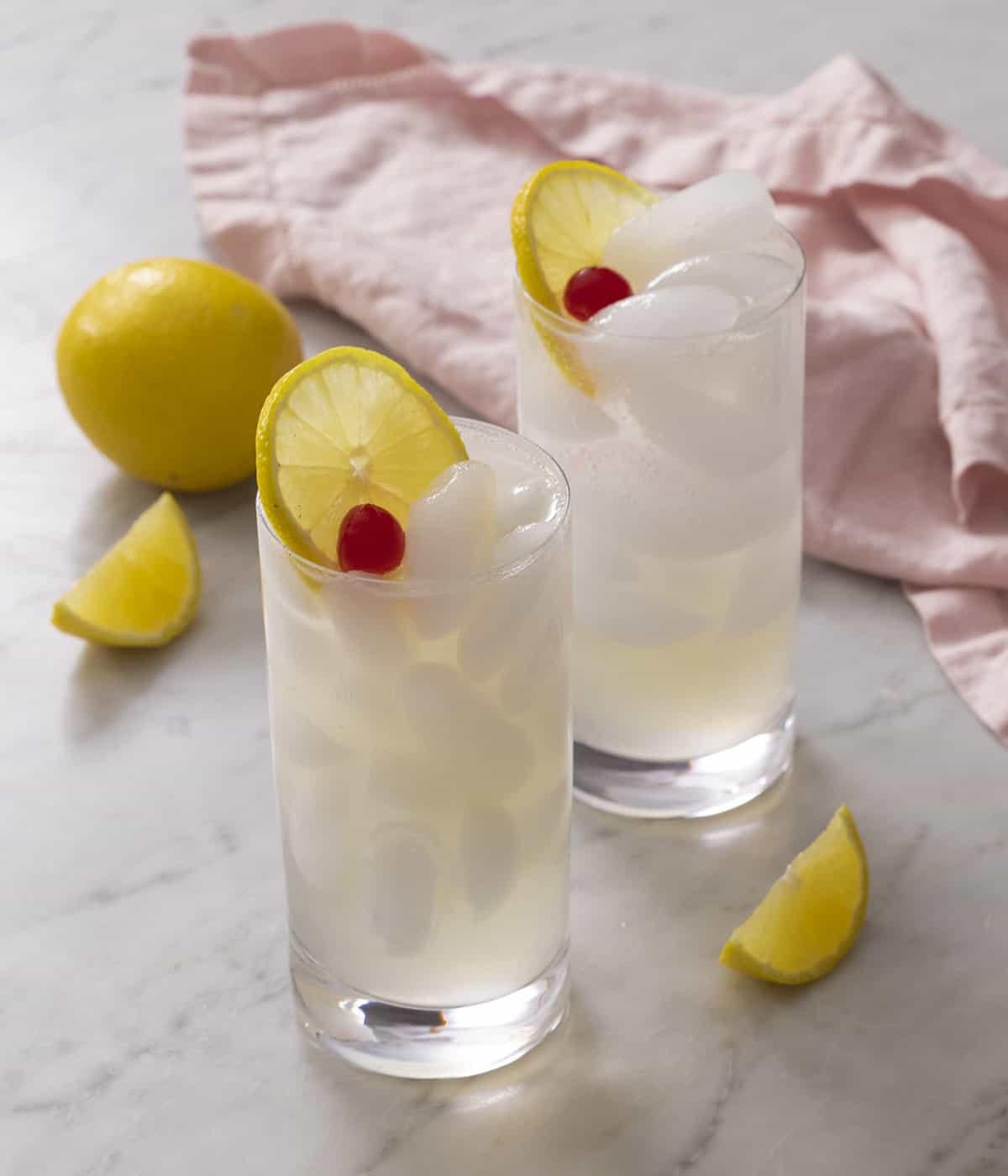 Two Tom Collins topped with cherries and lemon slices on a marble surface