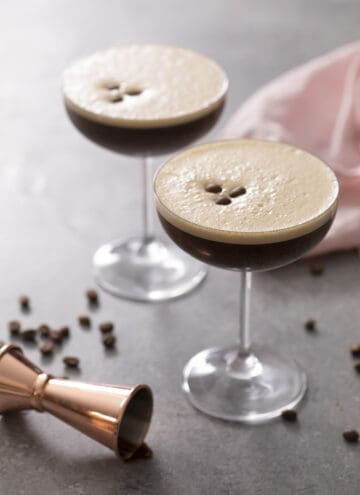 An espresso martini on a gray surface with coffee beans scattered around.
