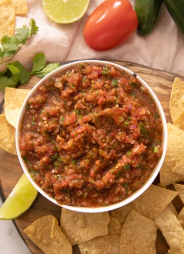 A close up of salsa in a bowl with tortilla chips and ingredients scattered around it.