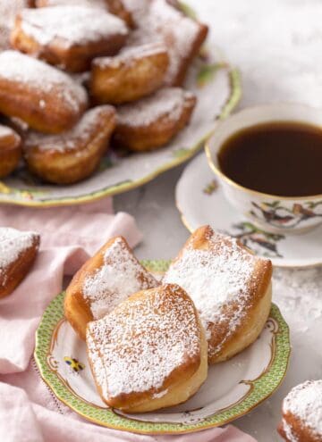 Three beignets on a plate with a cup of tea and platter of more beignets in the back.