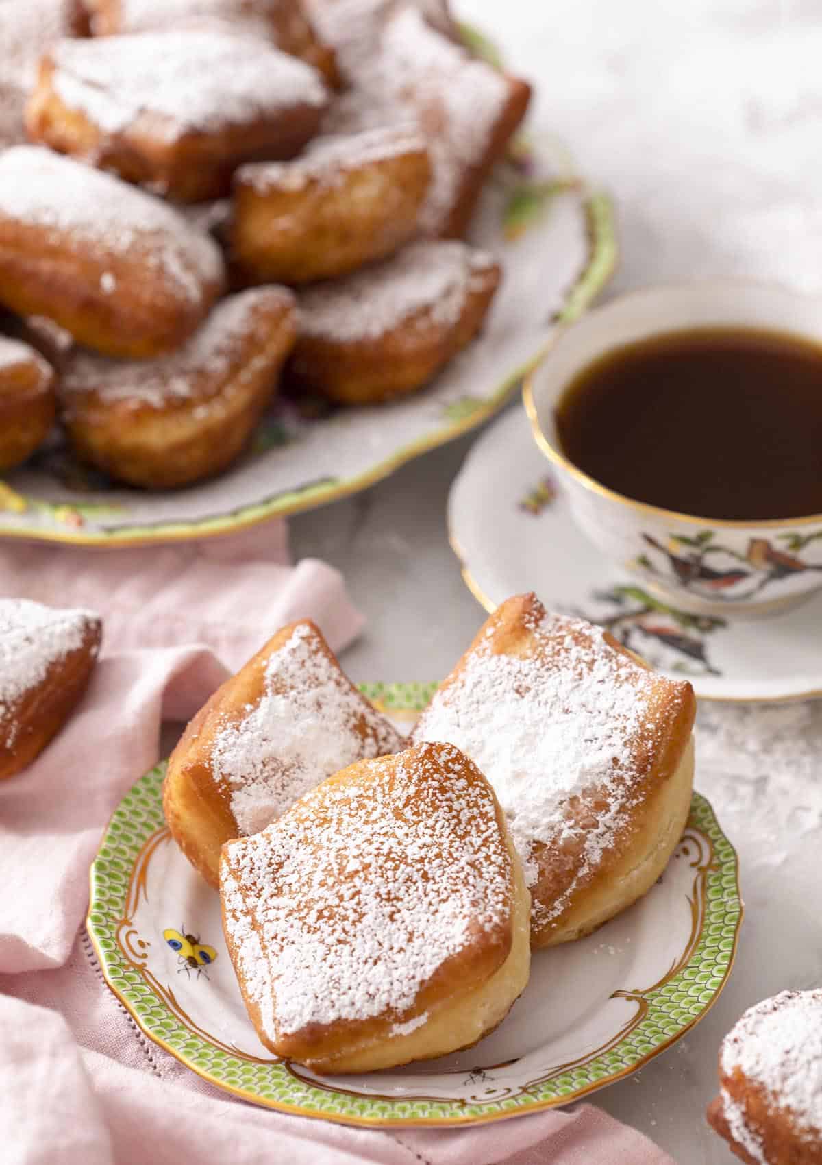 Three beignets on a plate with a cup of tea and platter of more beignets in the back.