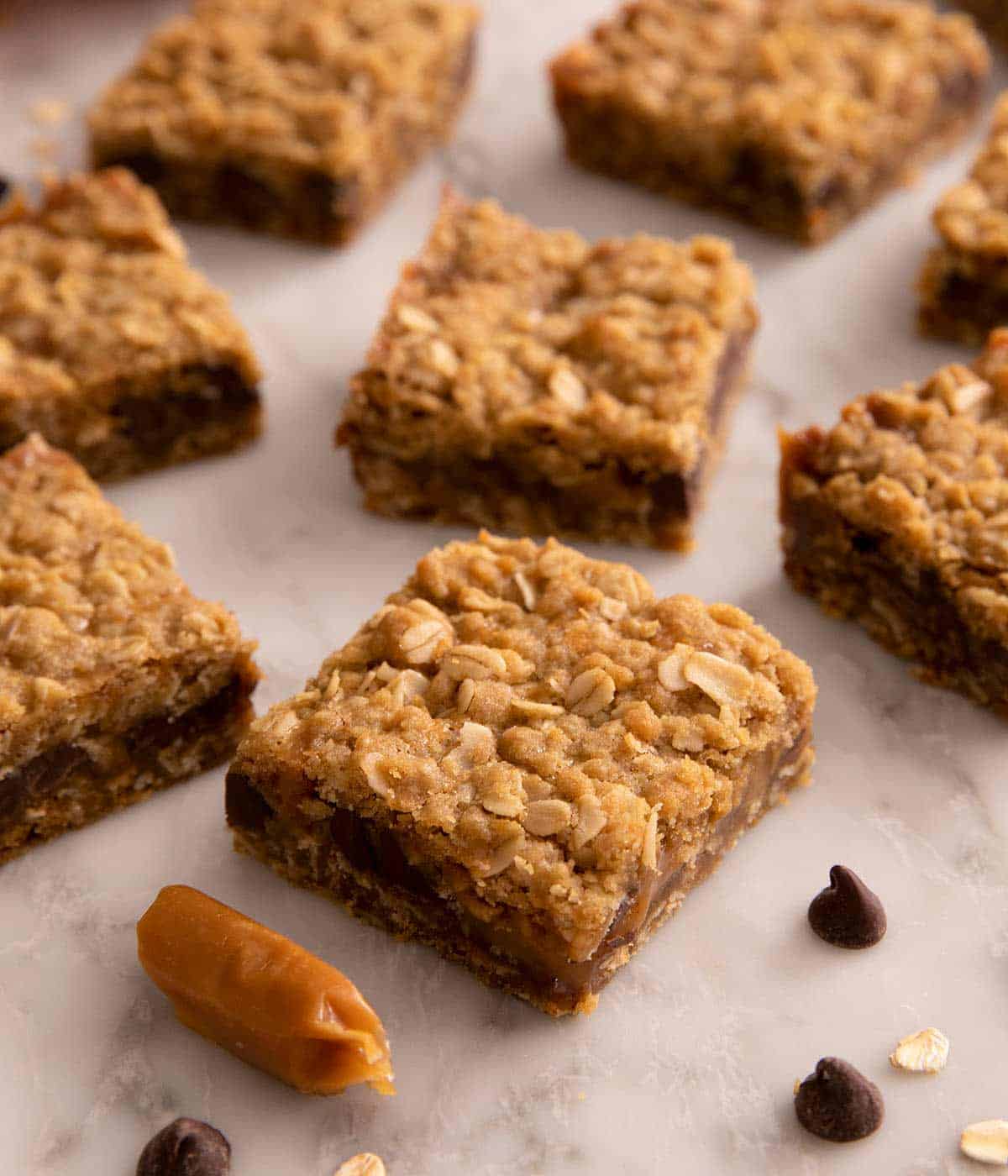 Carmetlita squares on a marble work surface. Chocolate chips, caramels, and oats scattered in front. 
