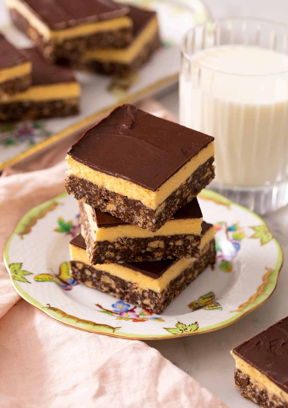 A stack of three Nanaimo bars on a plate. A glass of milk in the back.