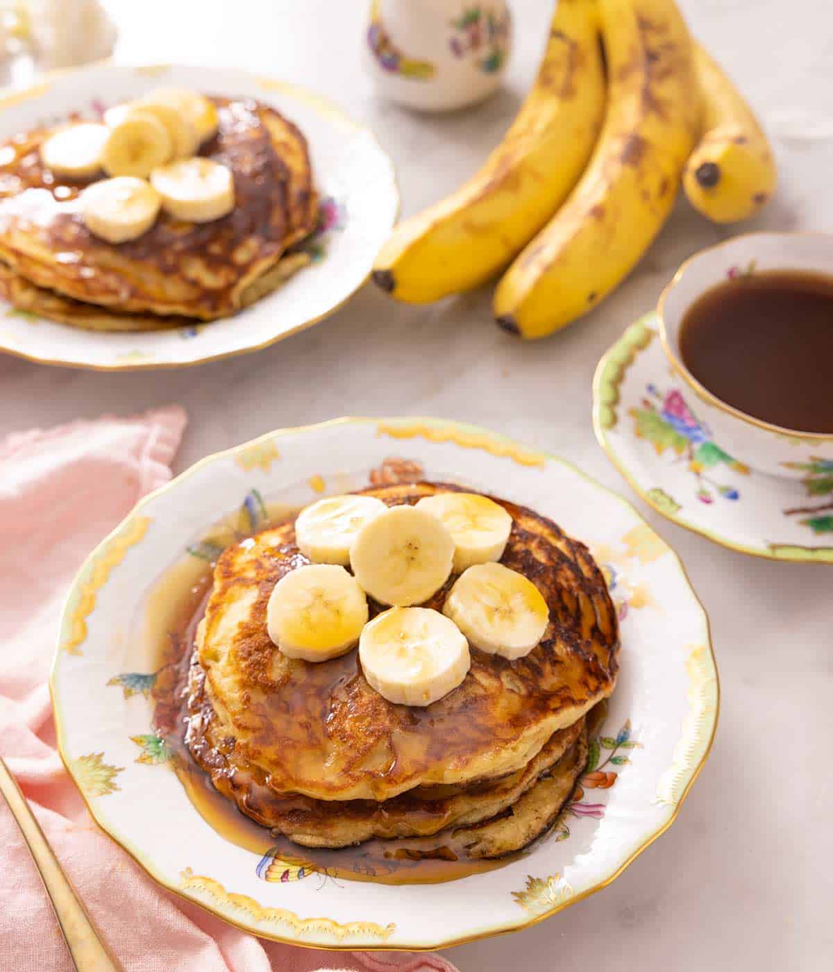 Banana pancakes on a plate topped with slices of banana. Coffee, bananas, and another plate of bananas in the background. 