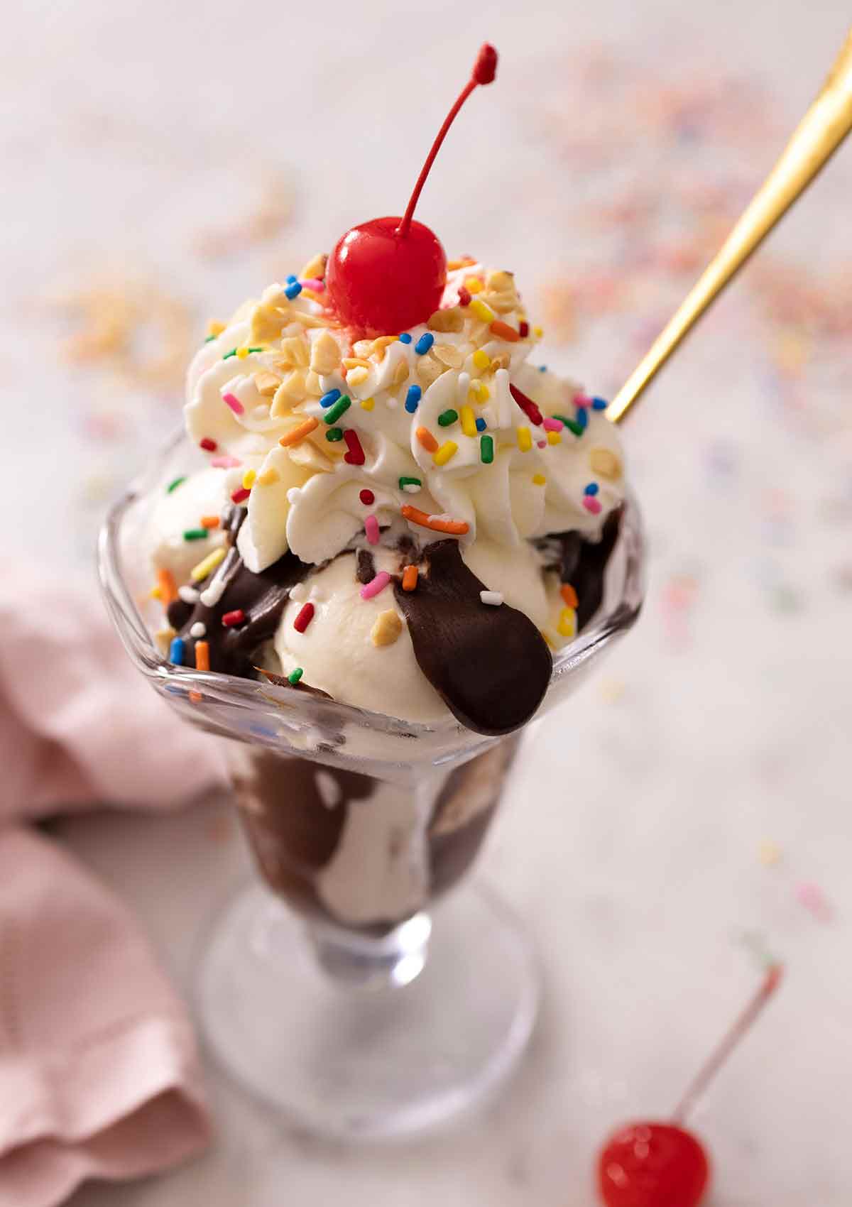 A glass of a hot fudge sundae made with vanilla ice cream with whipped cream, nuts, sprinkles, and a cherry on top.
