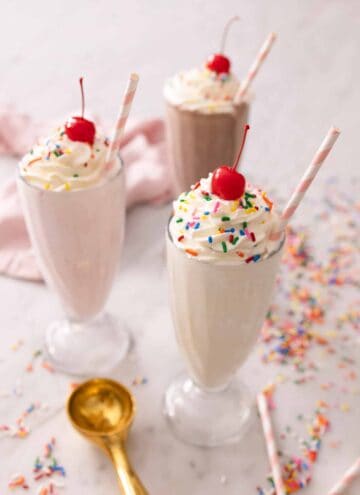 A vanilla milkshake, a strawberry milkshake, and a chocolate milkshake with toppings on top with sprinkles around the table.
