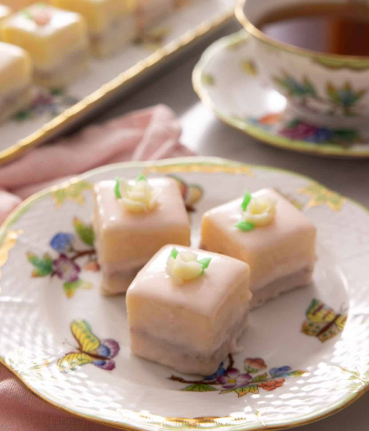 Three petit fours on a porcelain butterfly plate by a cup of tea.