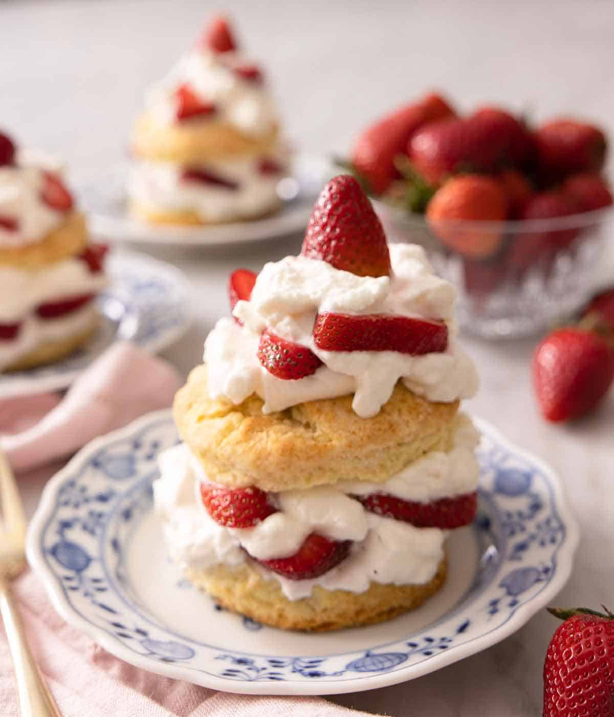 Three plates of strawberry shortcakes beside a bowl of strawberries with one plate in focus.