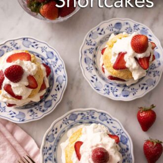 Pinterest graphic of an overhead view of three strawberry shortcakes by a bowl of strawberries. All cakes are topped with whipped cream and berries.