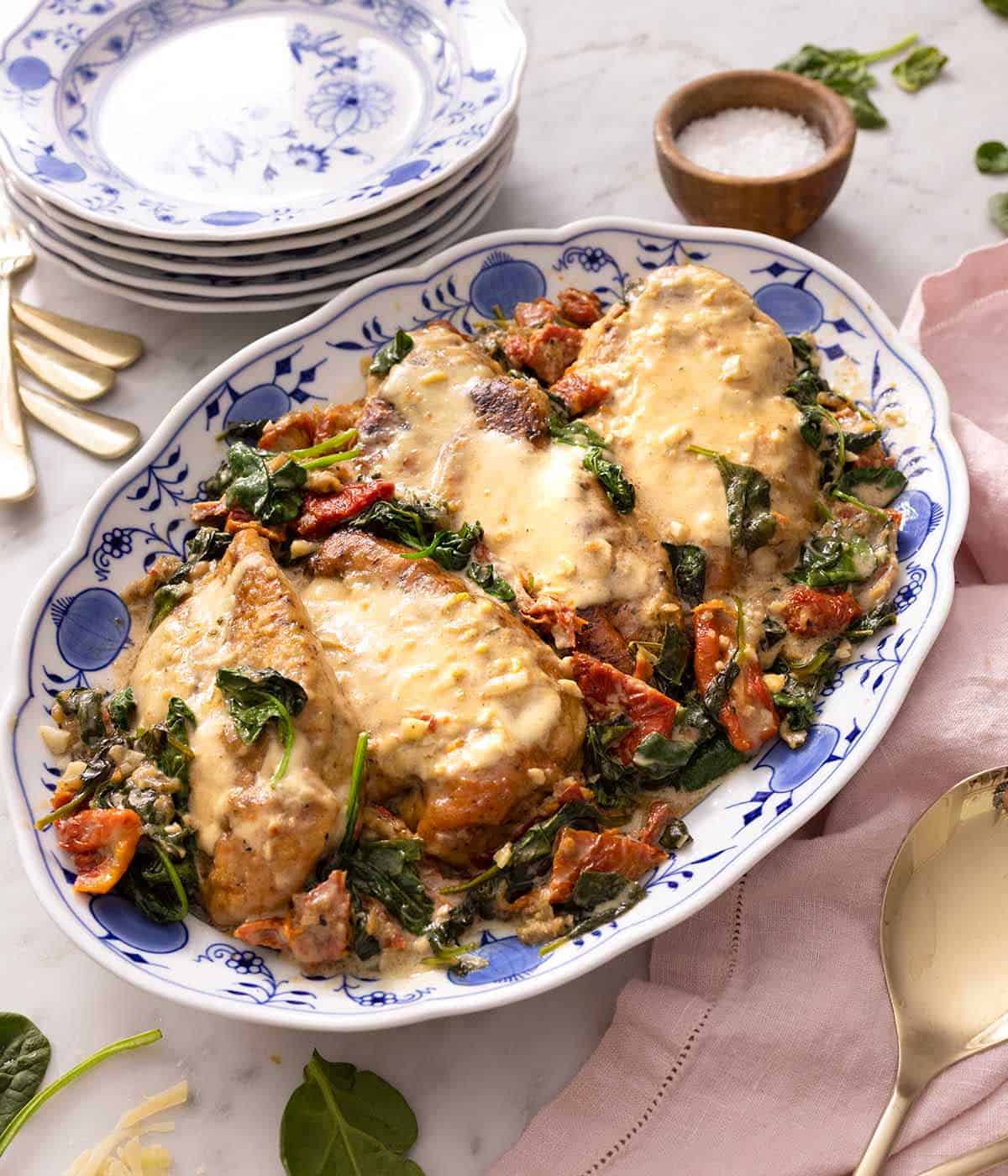 A white and blue floral platter with Tuscan chicken beside serving plates and a pink linen.
