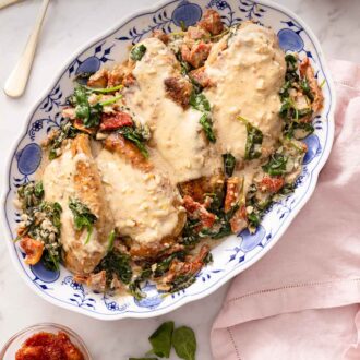 Pinterest graphic of a large blue and white porcelain platter with four servings of Tuscan chicken beside a pink linen, spinach, sundried tomatoes, and salt.