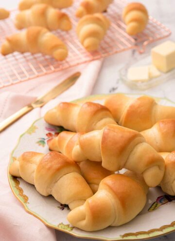 A plate of multiple crescent rolls with a cooling rack with more rolls in the background.