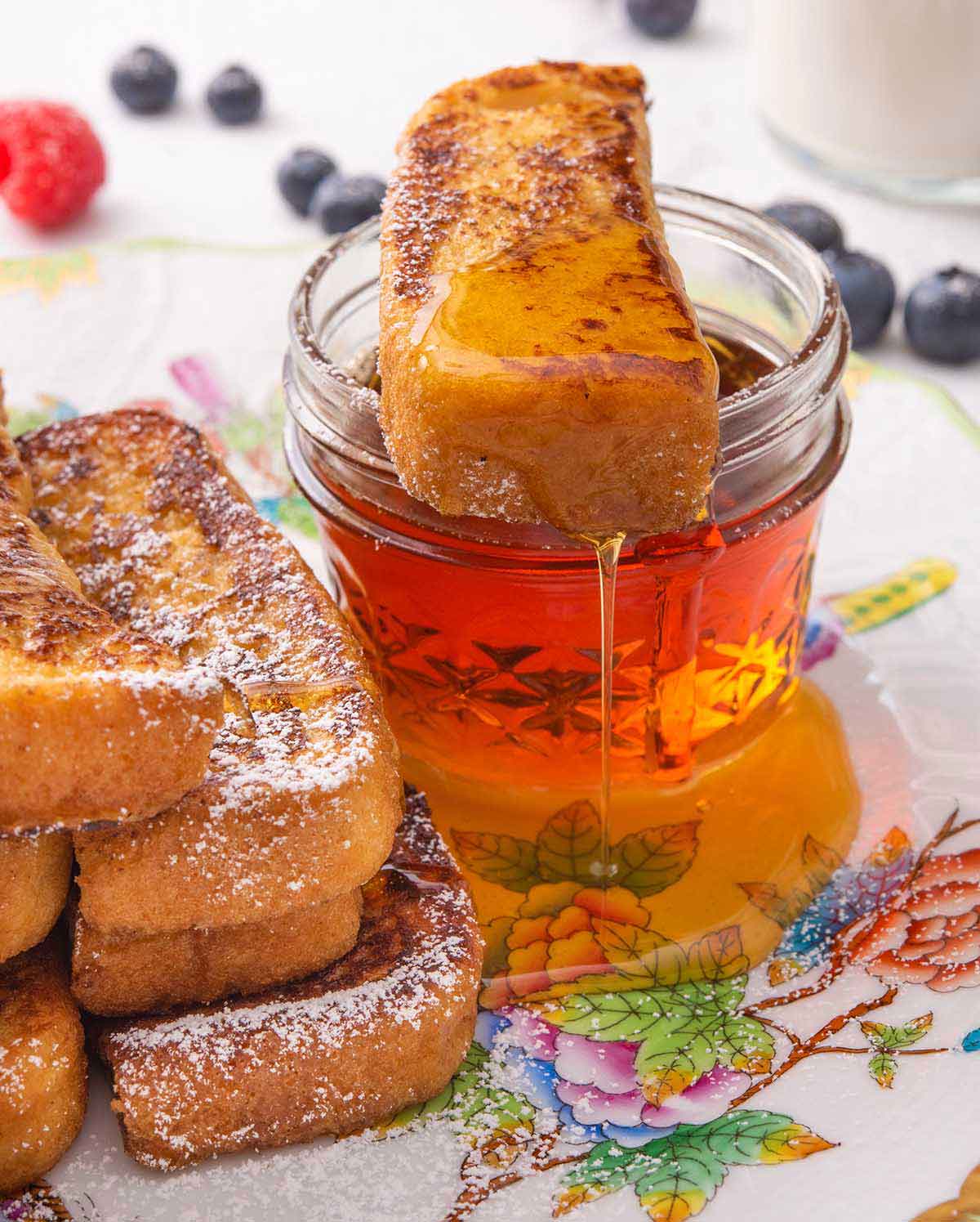 A plate with a French toast stick on top of a small mason jar of maple syrup with some dripping off the stick.