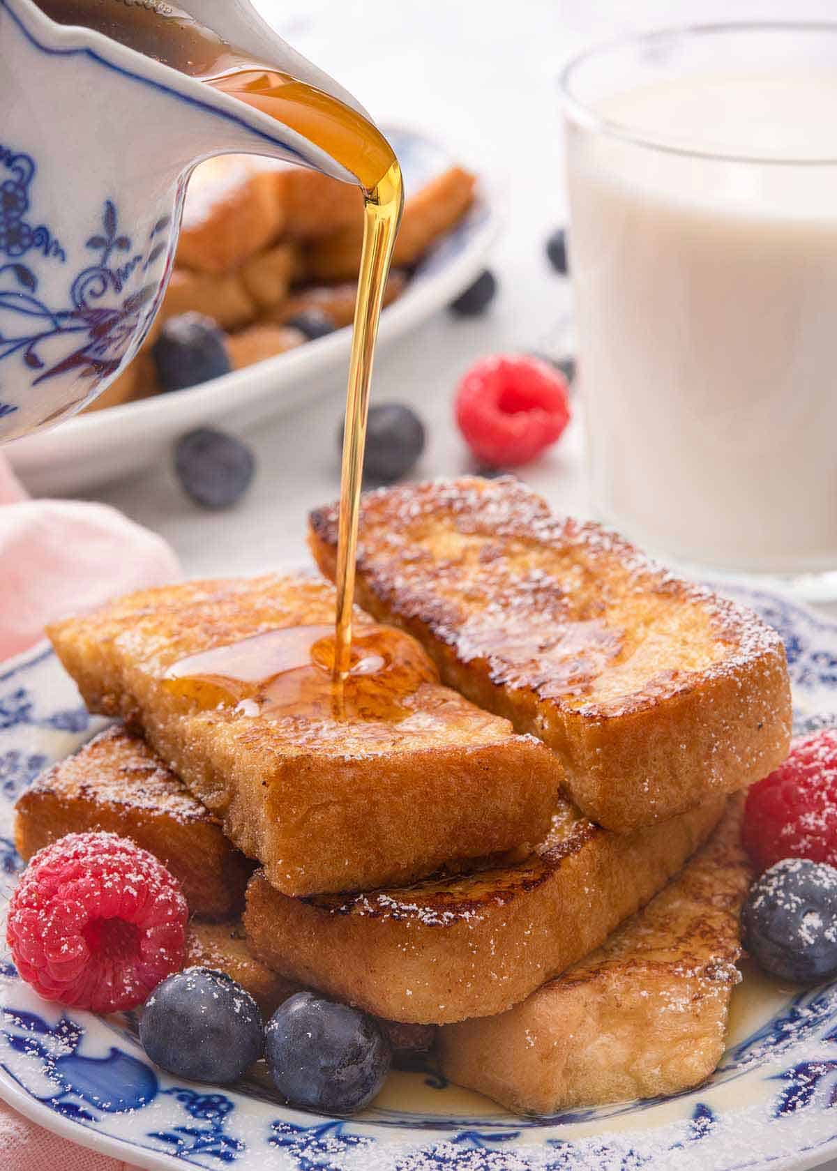 Maple syrup being poured on top of a stack of French toast sticks with fresh berries beside them.
