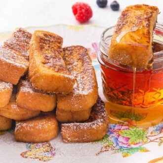 A plate with multiple French toast sticks with one on top of a mason jar of maple syrup.