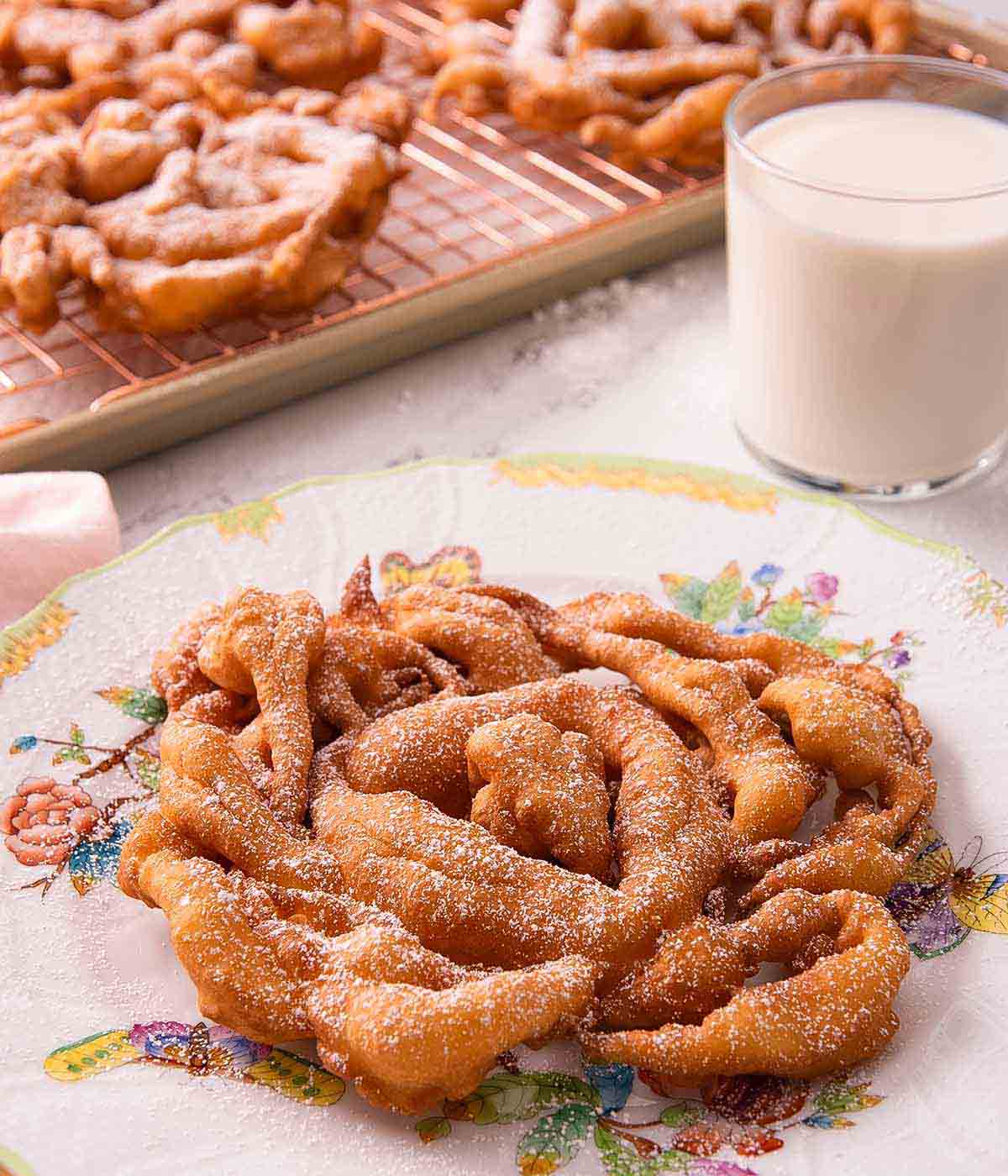 A round funnel cake with a fine dusting of powdered sugar with a glass of milk in the background and more funnel cakes on a cooling rack.