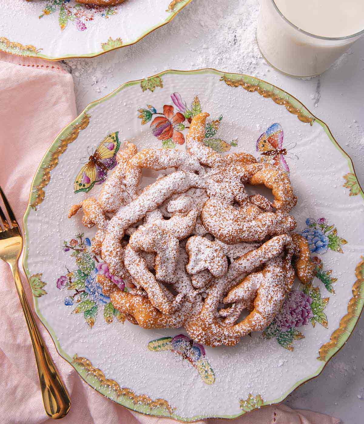 Overhead view of a powdered sugar covered funnel cake beside a gold colored fork and a pink linen napkin.