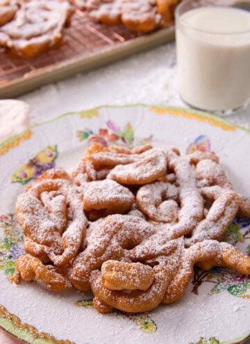 Funnel cake with a dusting of powdered sugar on a serving plate with a glass on milk in the background.