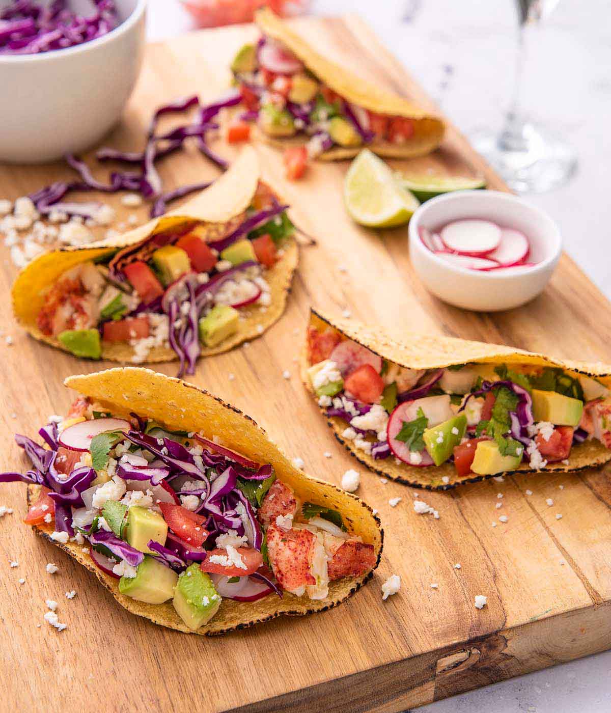 A brown cutting board with four lobster tacos with a pinch bowl of sliced radishes and a bowl of cabbage on the board.
