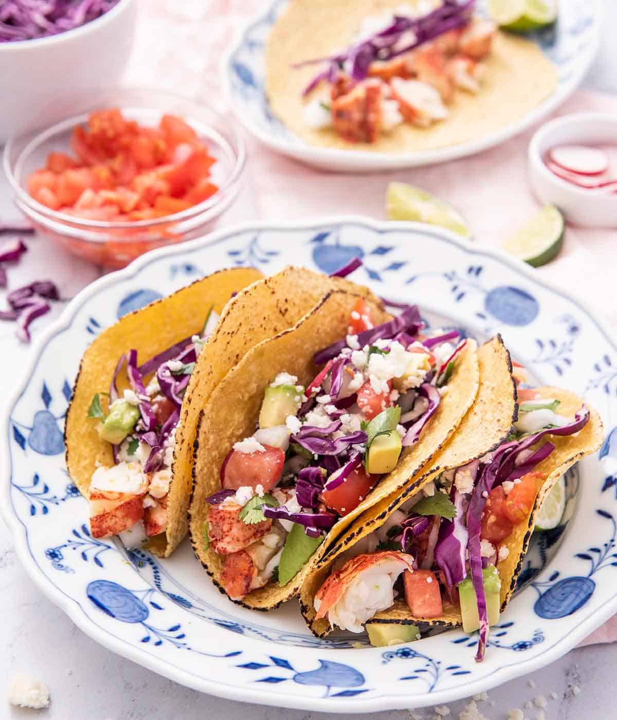 A blue and white plate holding three lobster tacos in a corn tortilla with ingredients in bowls in the background.
