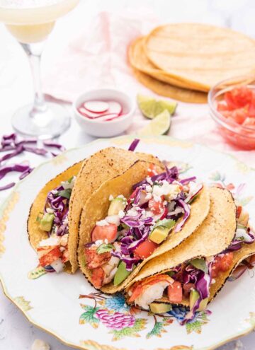 A plate of three lobster tacos in front of filling ingredients and a cocktail.