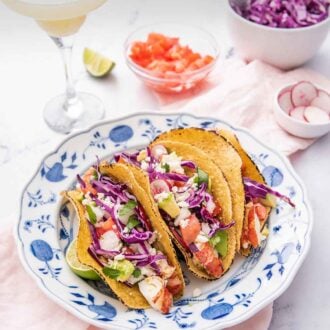Pinterest graphic of a blue and white plate with three lobster tacos beside a cocktail drink and three bowls of chopped tomatoes, radishes, and cabbage.