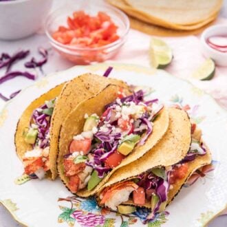 Pinterest graphic of three lobster tacos on a plate with crumbled cheese around it along with a bowl of tomatoes, cabbage, and tortillas in the background.