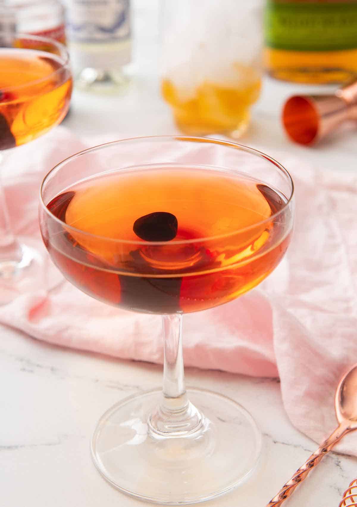 A Manhattan cocktail with a maraschino cherry in the middle in front of a pink linen napkin and the ingredients out of focus in the background.